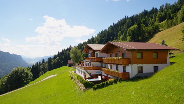 A two-story chalet with a balcony, surrounded by lush greenery and mountains.