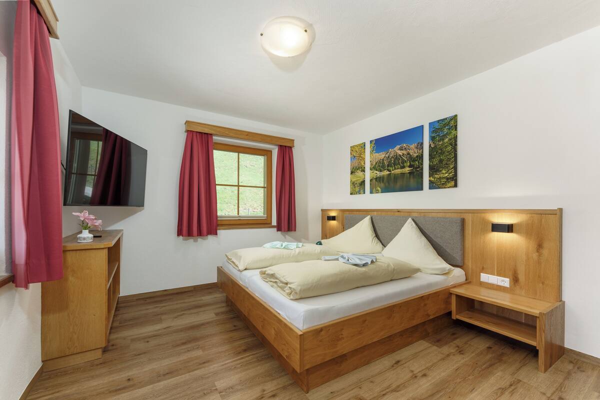 Une chambre à coucher avec un lit en bois, une table de chevet, une télévision et une fenêtre avec des rideaux.