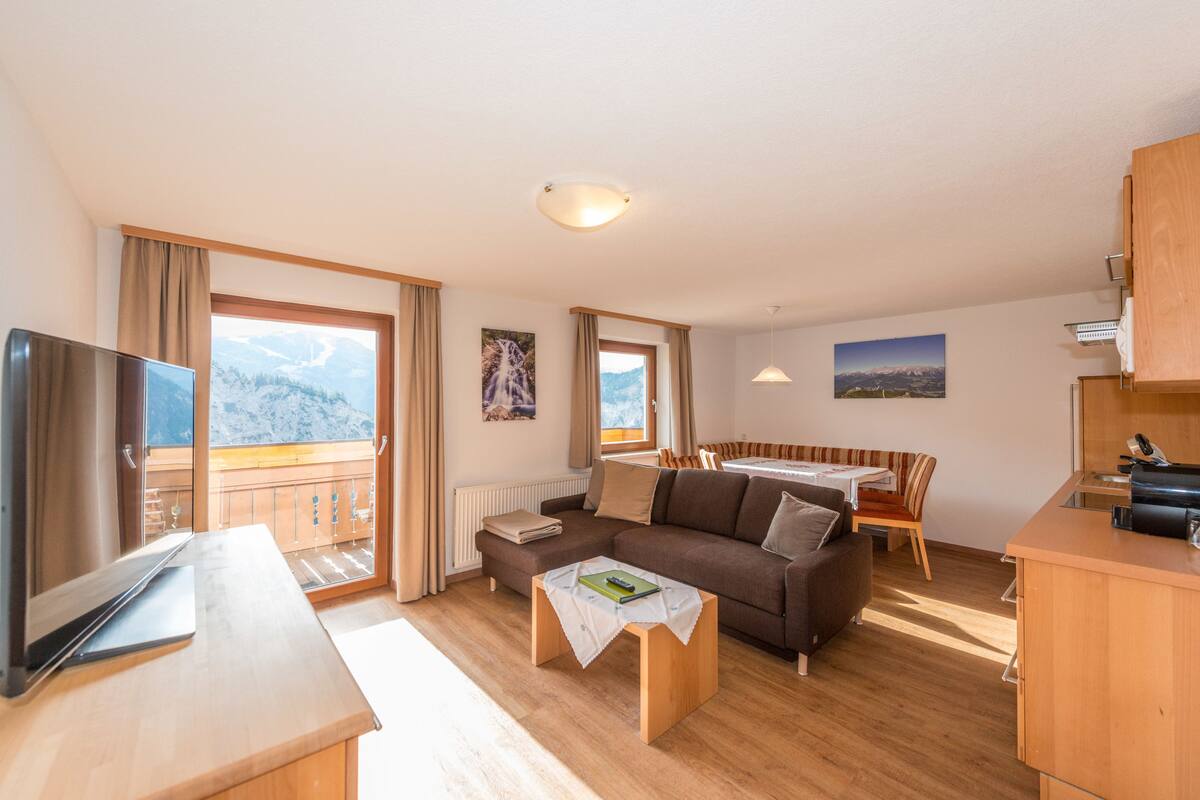 Un salon confortable avec un canapé, une table basse et un téléviseur. Il y a un balcon avec vue sur les montagnes.
