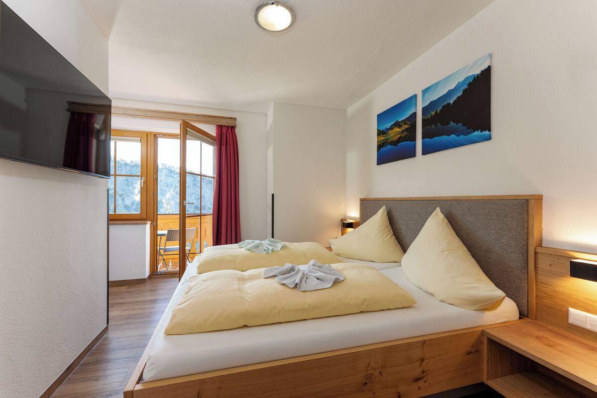 Une chambre d’hôtel avec un lit en bois, deux oreillers, une table de chevet, un téléviseur fixé au mur et une fenêtre donnant sur des arbres.