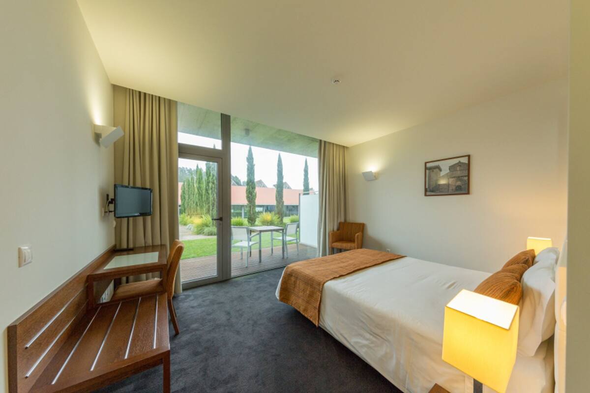 Ein Hotelzimmer mit Bett, Schreibtisch, Stuhl, Fernseher und einem großen Fenster mit Blick auf Bäume und Gebäude.
