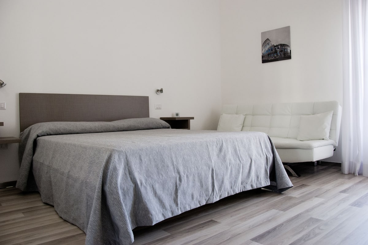 A modern hotel room with a large bed, a bedside table, a white sofa, and a painting on the wall.
