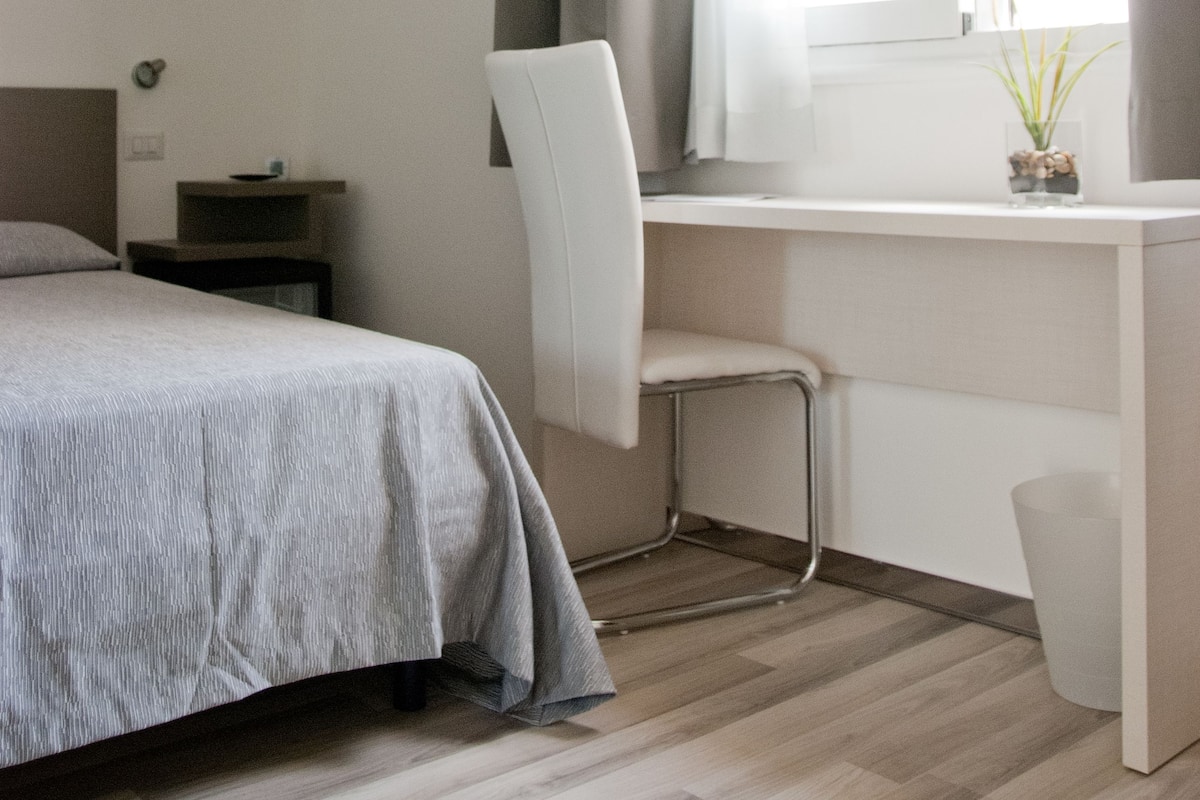 A bedroom with a bed, a desk, a chair, a window with curtains, and a small plant on the desk.