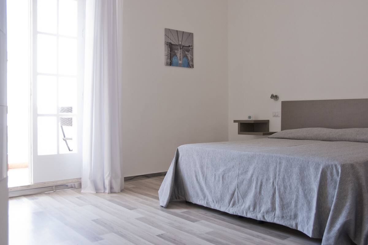 A bedroom with a bed, a window with curtains, a wall-mounted shelf, and a picture on the wall.