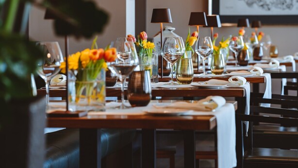 Une salle de restaurant avec des tables dressées pour le service, agrémentée de vases remplis de fleurs et de verres à vin.