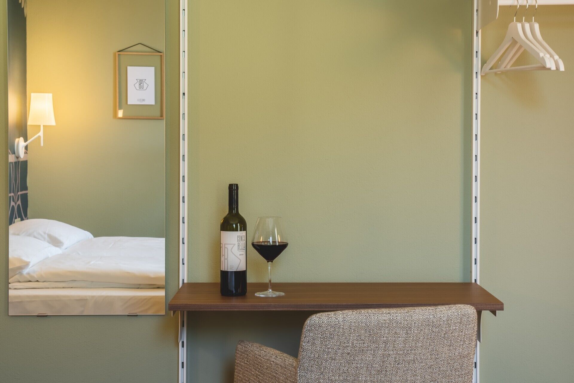 A hotel room with a bed, a small table with a bottle of wine and a glass, a lamp, and a wall-mounted picture.
