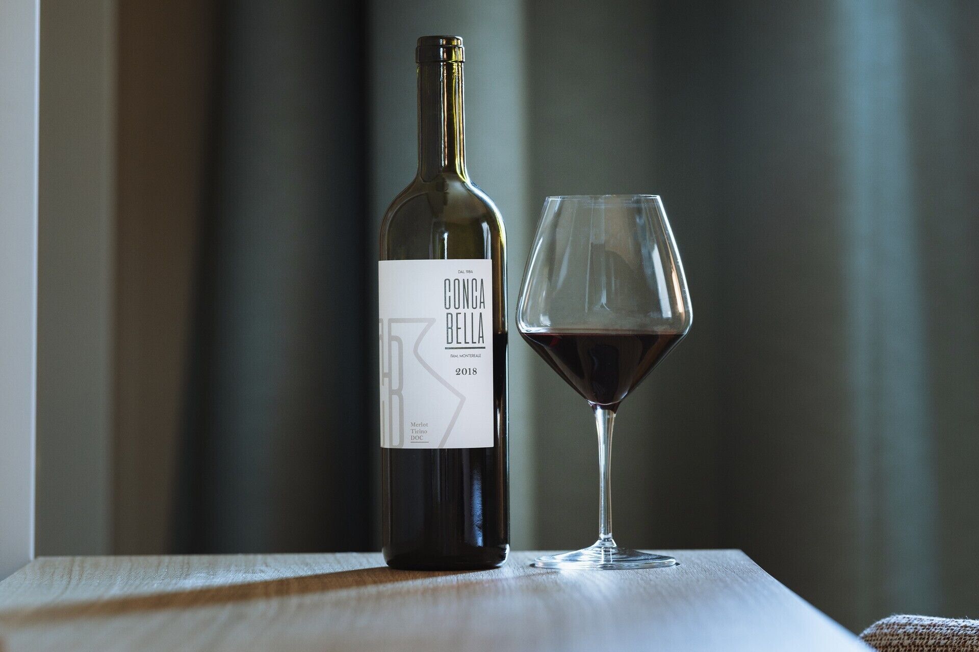 A bottle of Conca Bella Merlot 2018 wine and a glass of wine on a wooden surface.