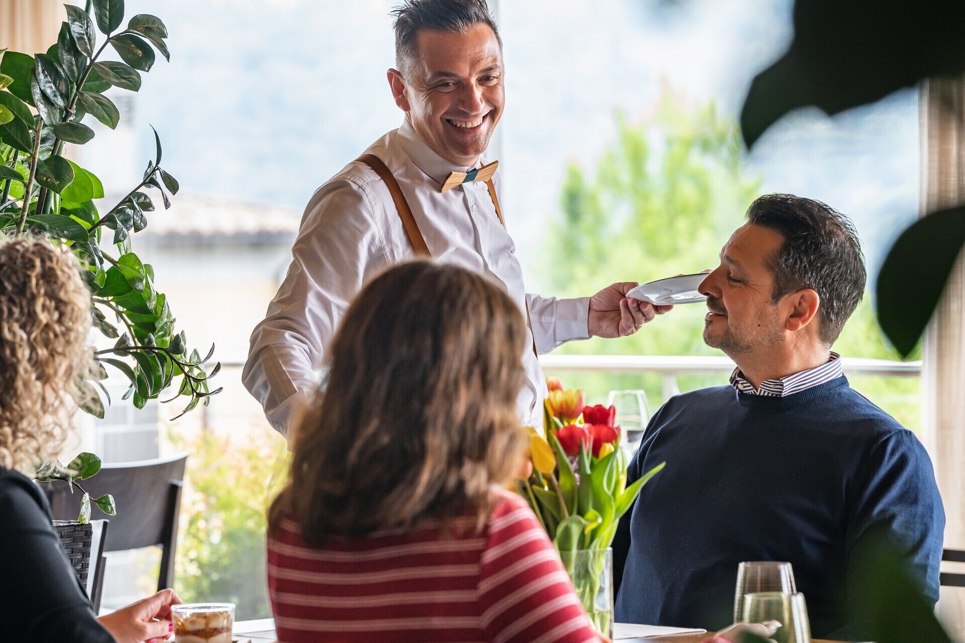 A waiter serving a customer at a table with a view of greenery and a body of water.