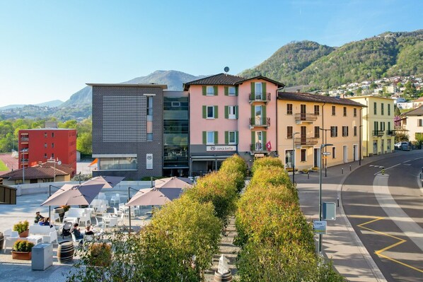 A hotel with a modern facade, a visible restaurant with outdoor seating, and a mountain backdrop.