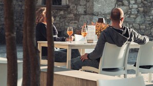 Un couple est assis à une table en plein air, devant un menu, avec deux verres de vin, et un mur de pierres en arrière-plan.