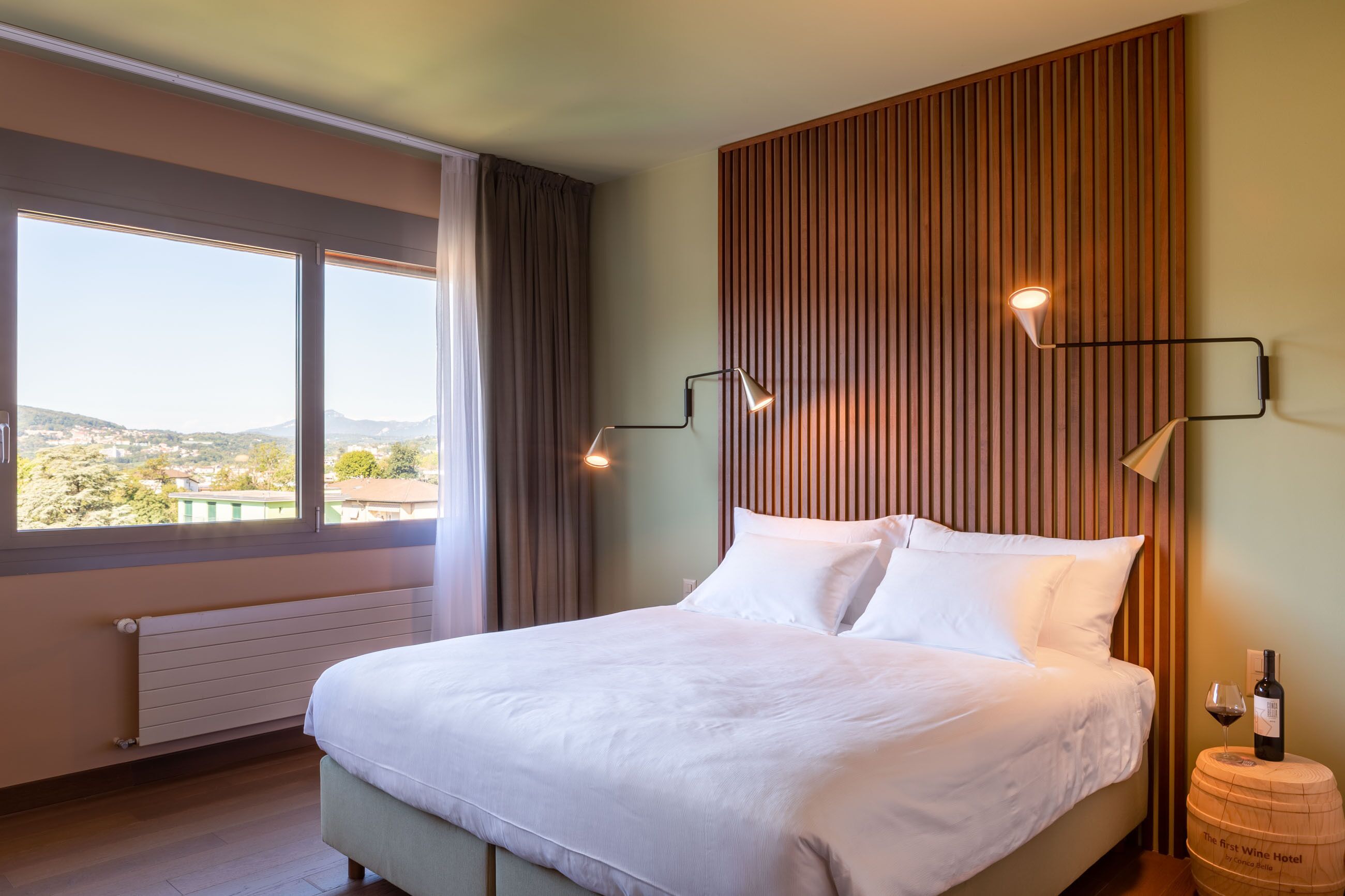 Een hotelkamer met een groot bed, twee bedlampen, een wand van houten latten en uitzicht op het landschap.