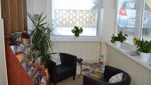 A cozy reading nook with a black armchair, a glass table with magazines, a potted plant, and a window with blinds. - Pension Nika (Prague)