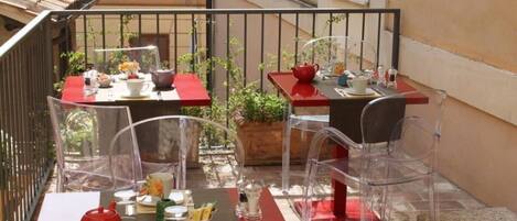 A balcony with two tables, each with a red tablecloth and clear chairs. There are potted plants and a red teapot on the tables.