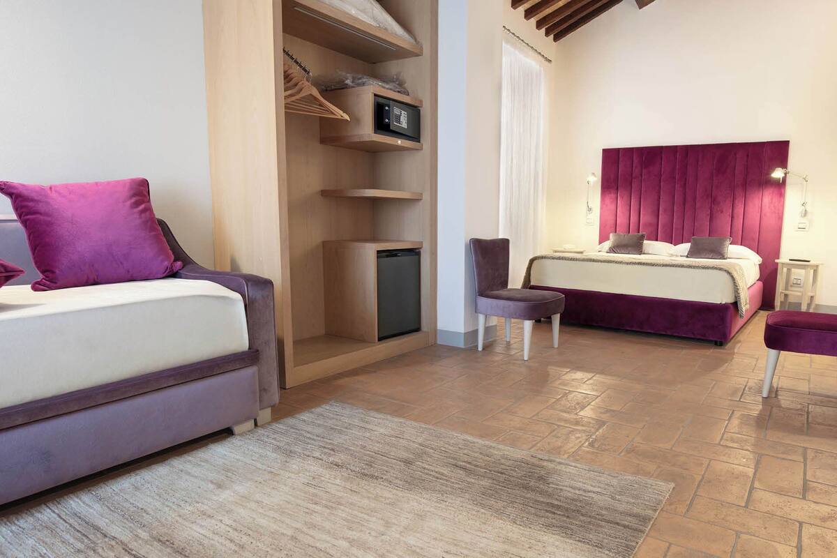 A bedroom with a bed, a chair, a small table, a shelf with a microwave, and a wardrobe.