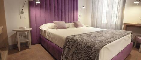 A bedroom with a large bed, a nightstand, a lamp, and a window with sheer curtains.