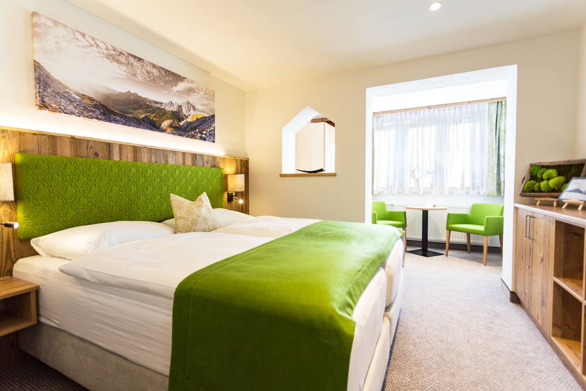 A hotel room with a large bed, a green headboard, a painting on the wall, a mirror, a window with curtains, and a small table with chairs.