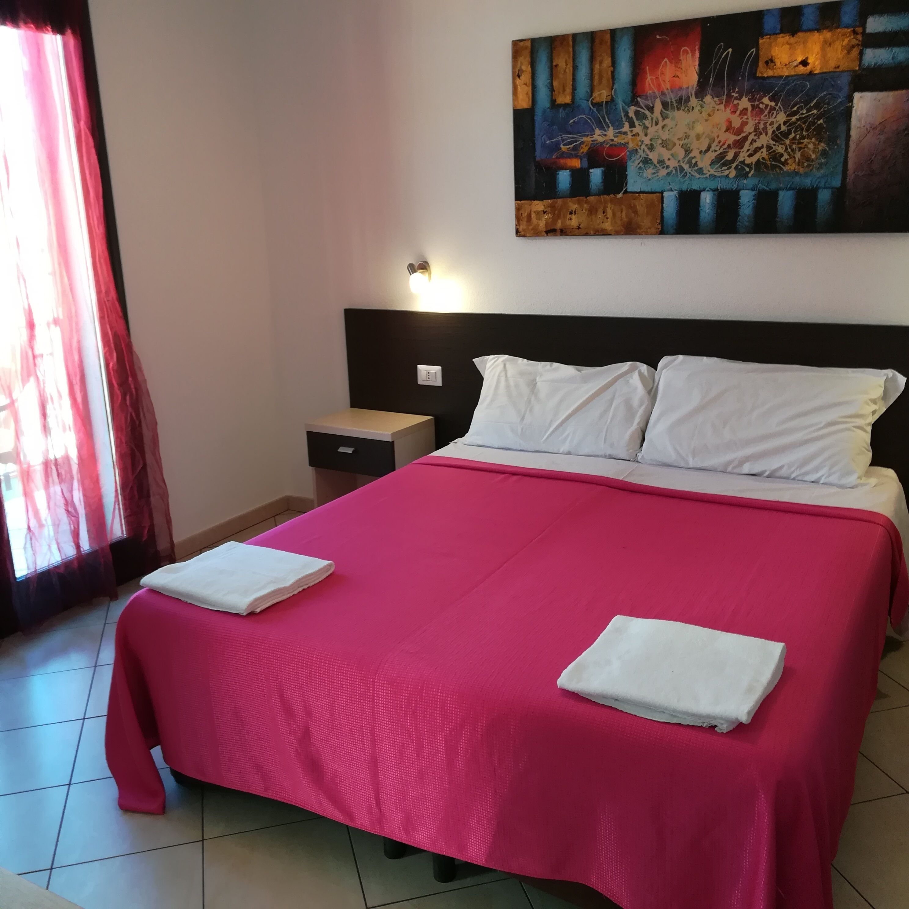 A hotel room with a large bed, a nightstand, a wall-mounted light, and a painting above the bed.