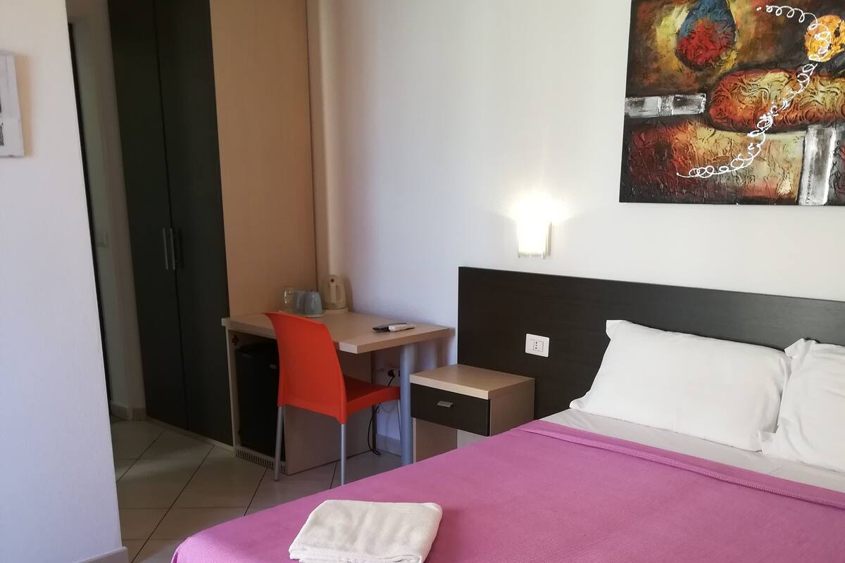 A hotel room with a bed, a desk, a chair, and a painting on the wall.