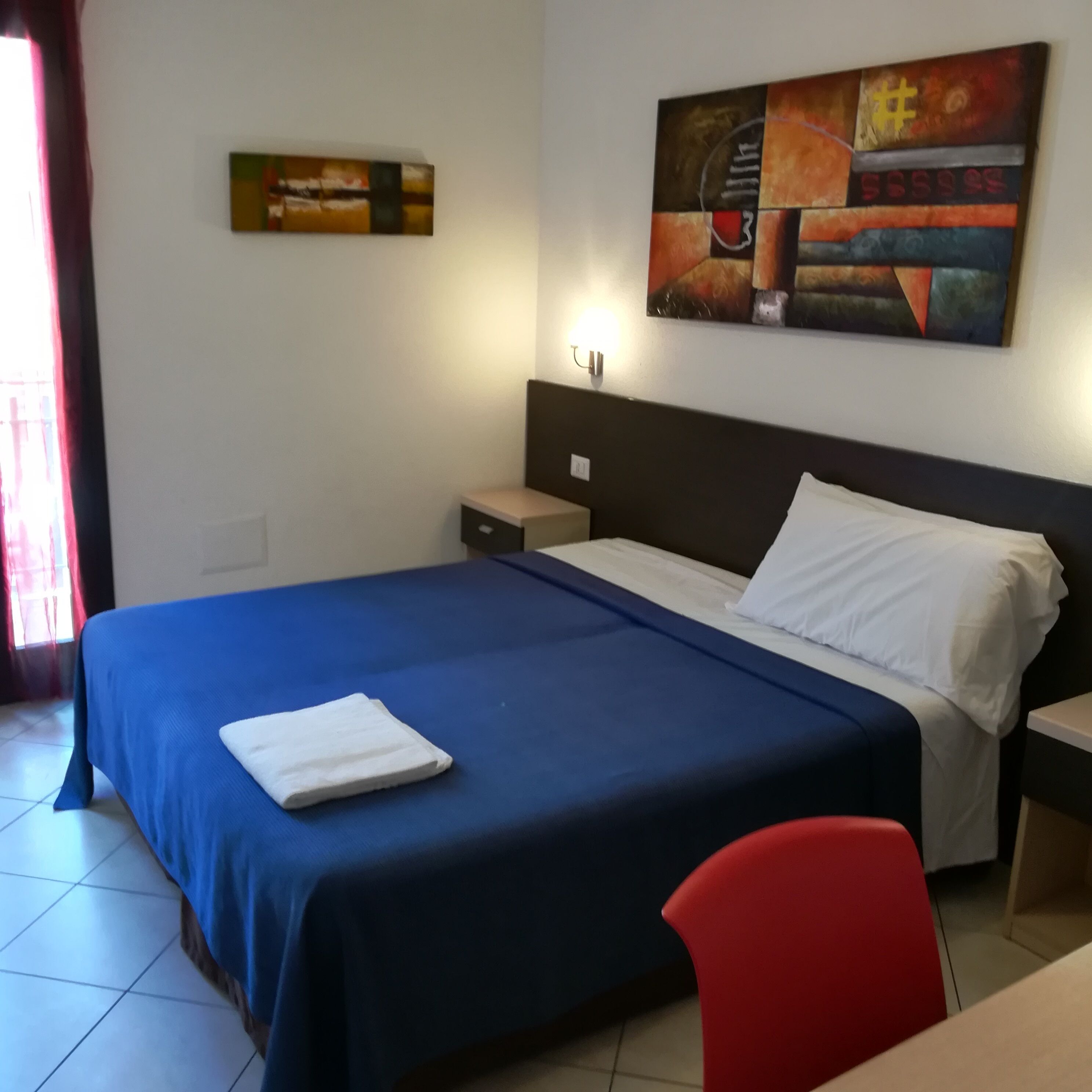 A hotel room with a large bed, a nightstand, a desk, and a chair. There is a painting on the wall and a window with curtains.