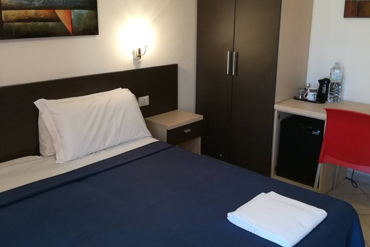 A hotel room with a bed, a desk, a wardrobe, and a painting on the wall.