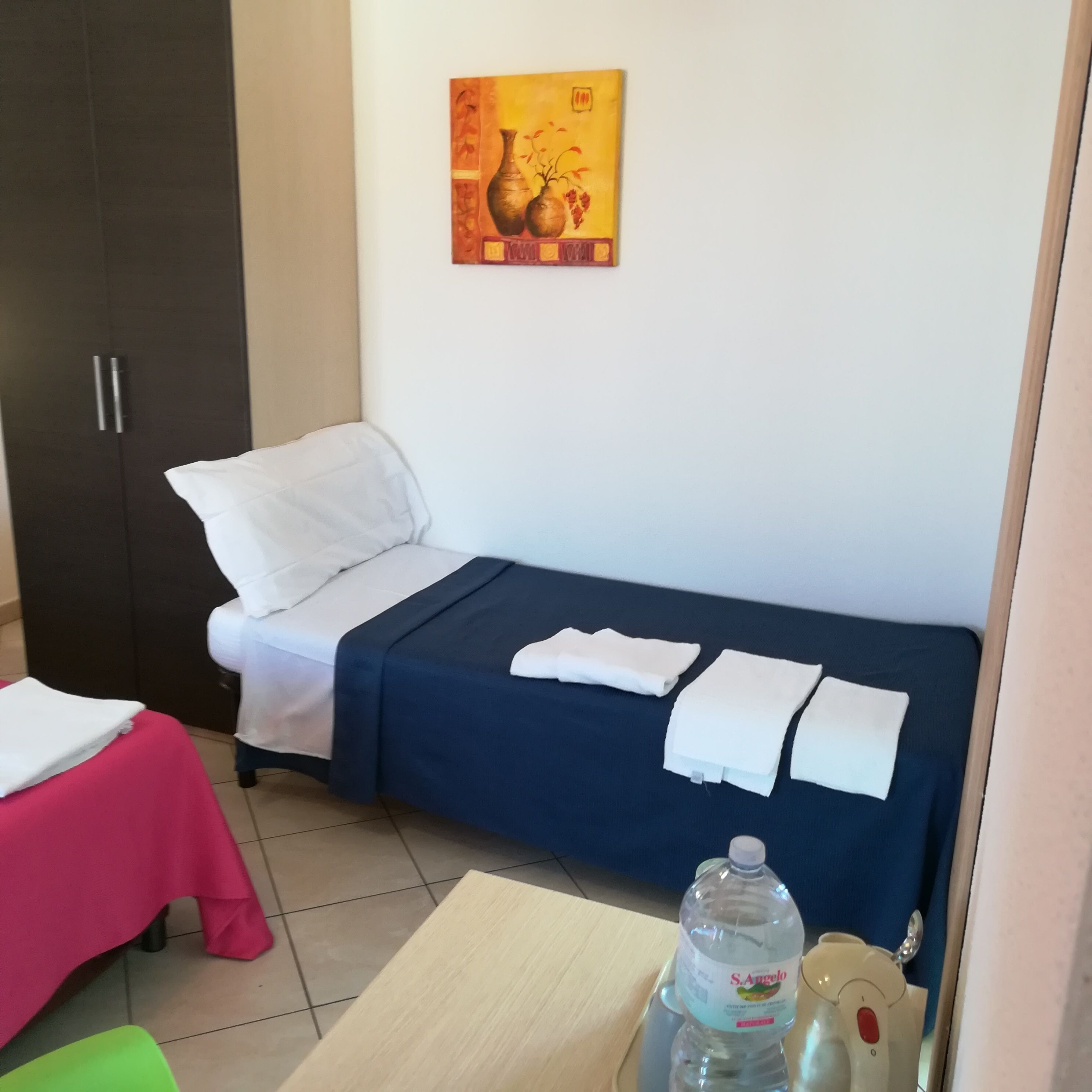 A small, neatly arranged room with a bed, a table with a bottle, and a painting on the wall.