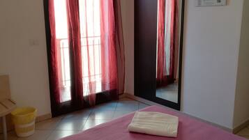 A hotel room with a bed, a mirror, a window with red curtains, a yellow trash bin, and a wall-mounted newspaper.