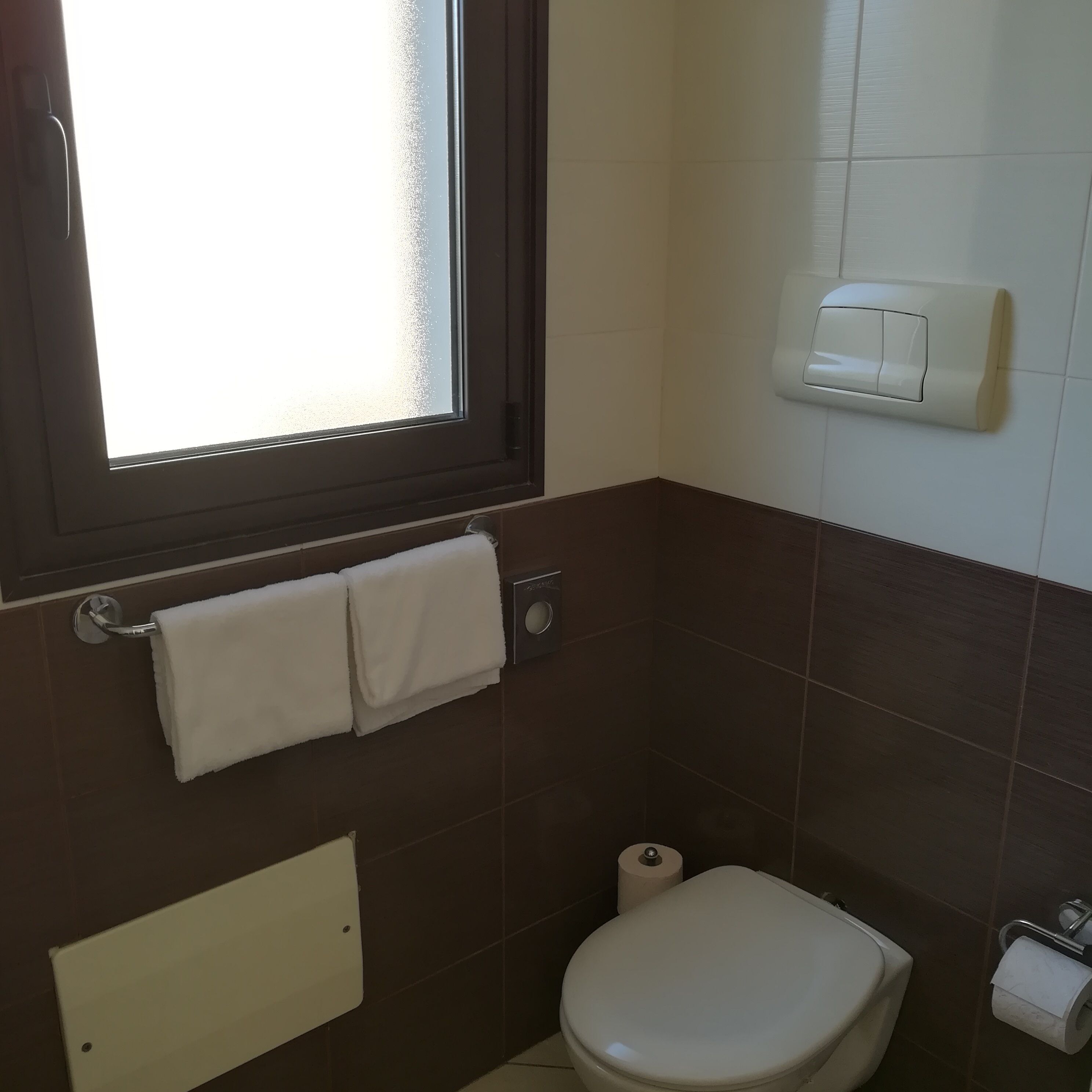 A small bathroom with a toilet, a towel rack with two towels, a window, and a paper towel dispenser.