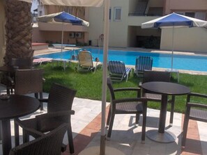 A poolside area with lounge chairs and umbrellas, a stone pillar, and a view of a swimming pool. - Mythos Beach Hotel (Platanias)