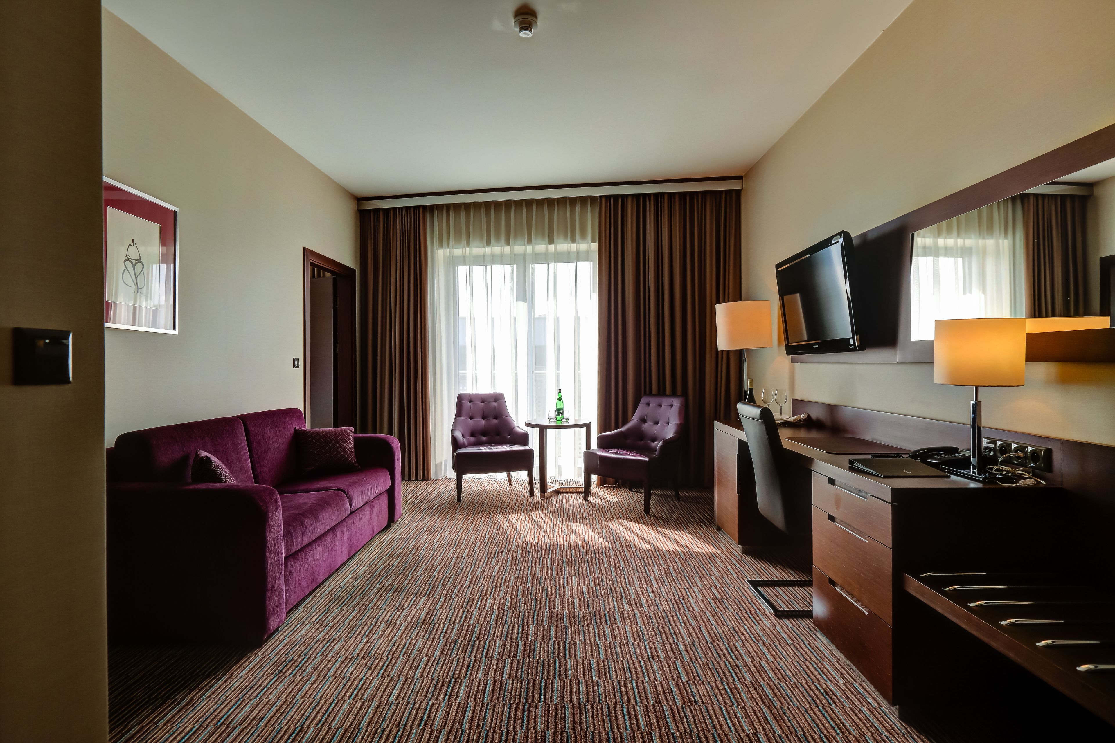 Ein Hotelzimmer mit einer Couch, zwei Sesseln, einem kleinen Tisch, einem Schreibtisch mit Fernseher und Teppichboden.