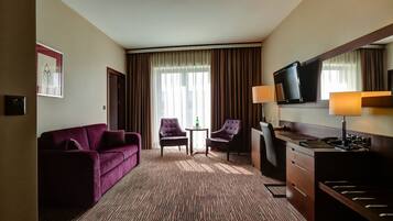 Ein Hotelzimmer mit einer Couch, zwei Sesseln, einem kleinen Tisch, einem Schreibtisch mit Fernseher und Teppichboden.