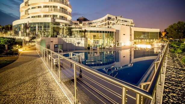 A modern hotel with a large pool, multiple floors, and a well-lit exterior.