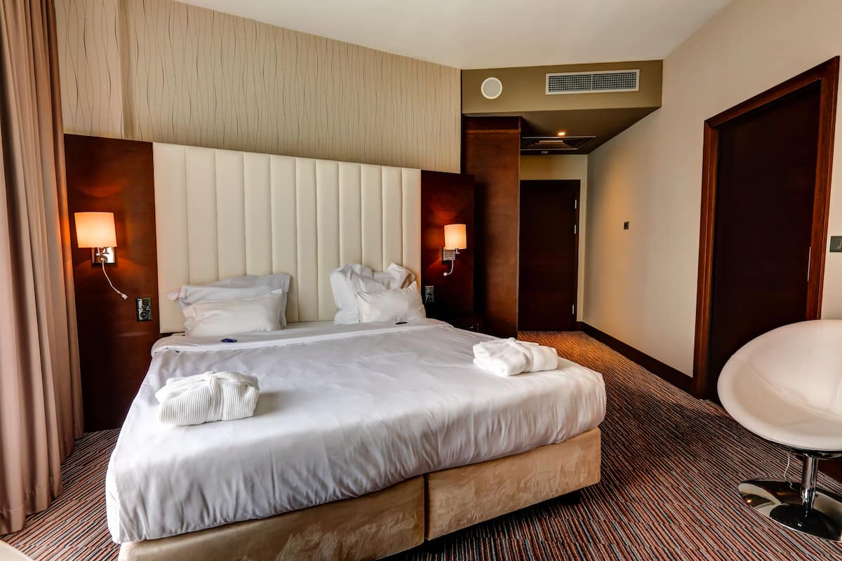 Quarto de hotel com uma cama grande, duas luminárias de cabeceira, um espelho redondo e uma porta para outro cômodo.