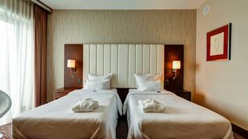 Quarto de hotel com duas camas, cabeceira, luminárias laterais, um quadro emoldurado e uma cadeira.