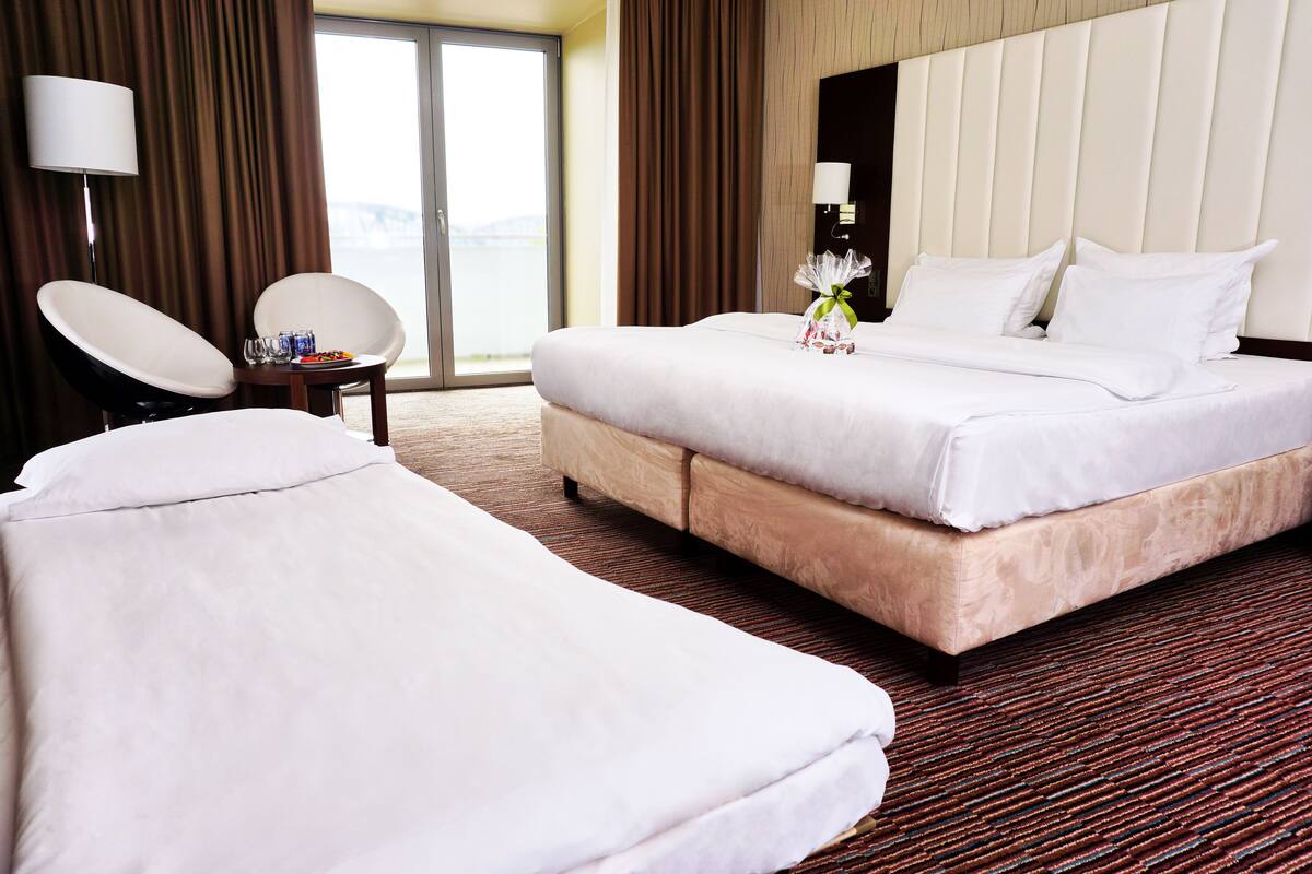 Quarto de hotel com uma cama grande, uma cama menor, uma cadeira, uma mesa com bebidas e vista para o exterior.