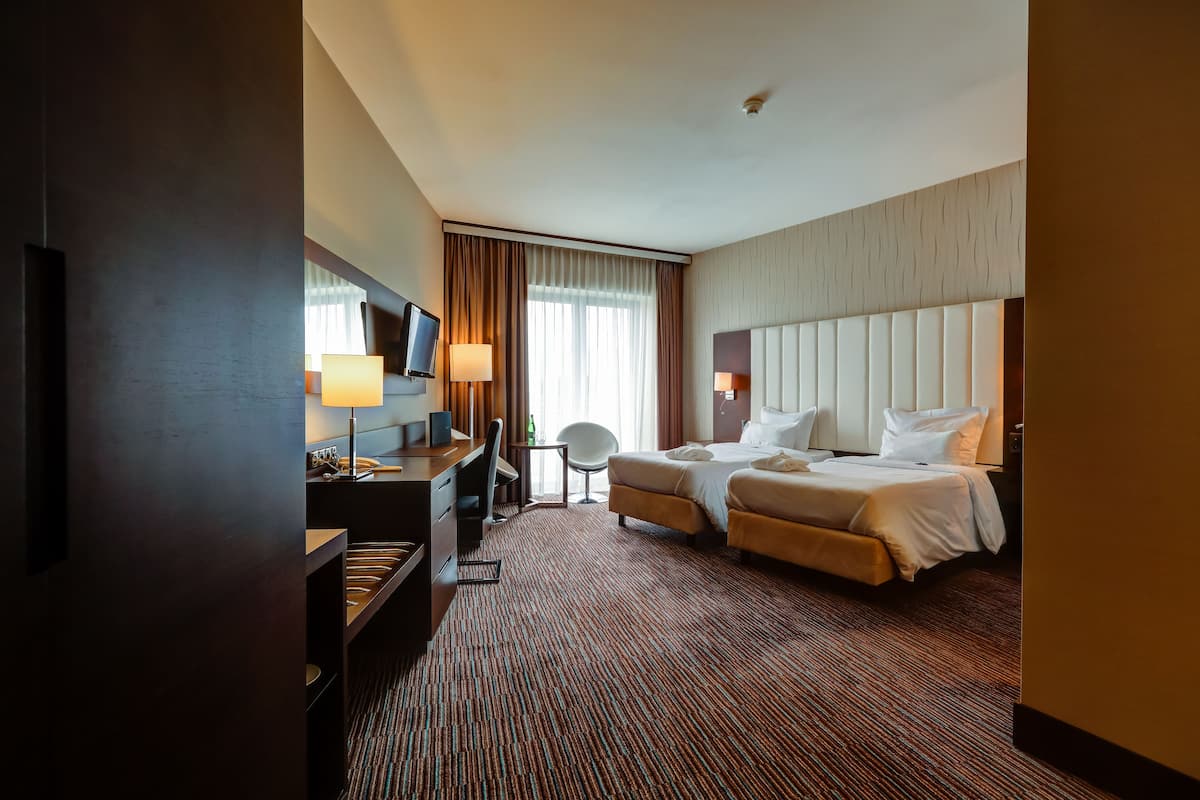 Quarto de hotel com cama, escrivaninha e cadeira, televisão e janela com cortinas.