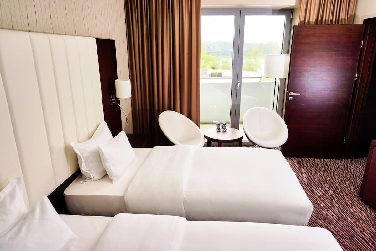 Quarto de hotel com uma cama, duas cadeiras brancas, uma mesa pequena, um abajur e vista para áreas verdes pela janela.