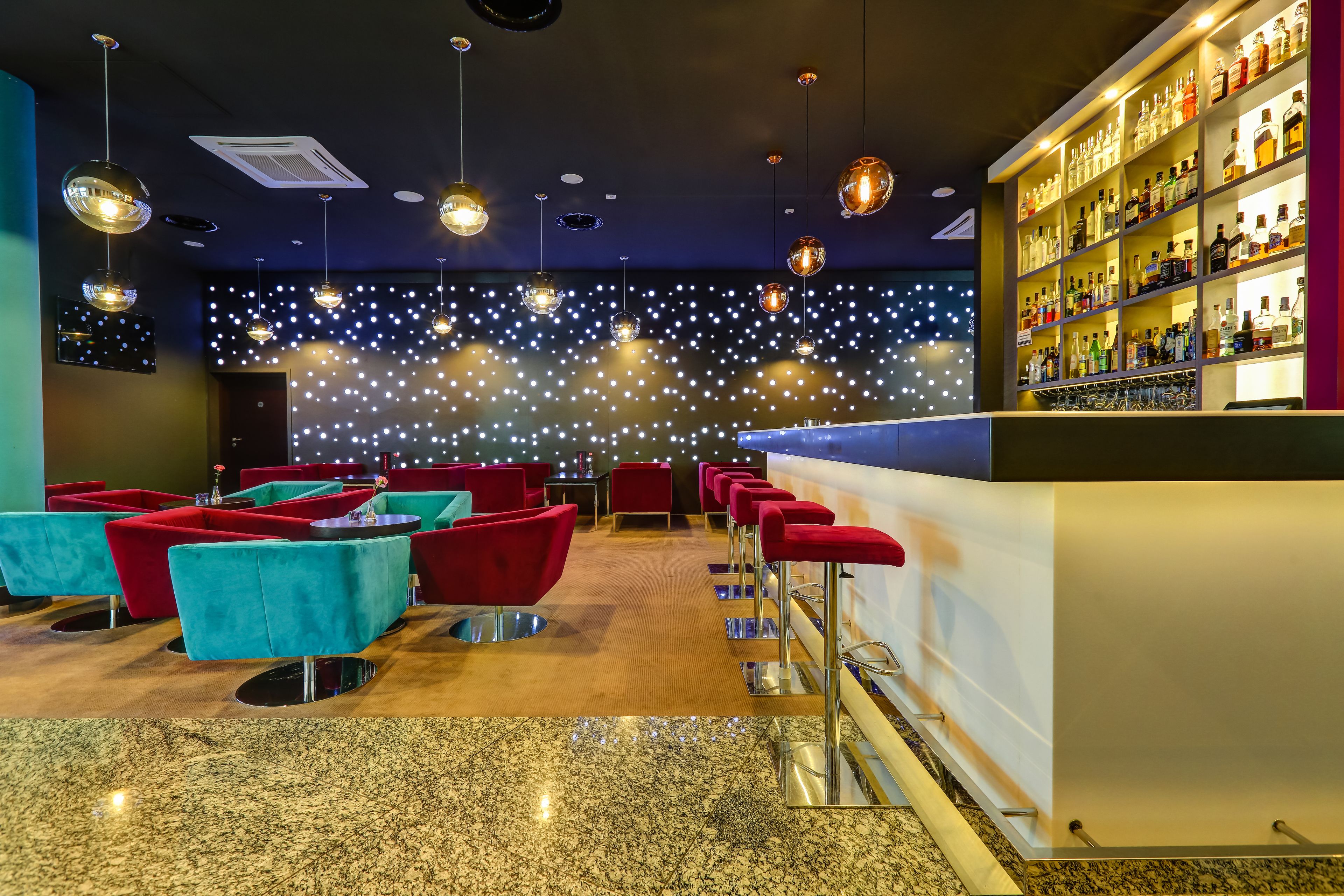 Un espace bar doté d’un sol en granit poli, de sièges rouges et turquoise, et d’un mur décoré d’étoiles illuminées.
