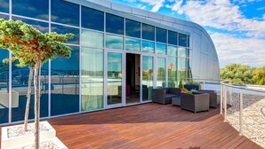 A modern building with large glass windows, a wooden deck, outdoor seating, and a small tree.