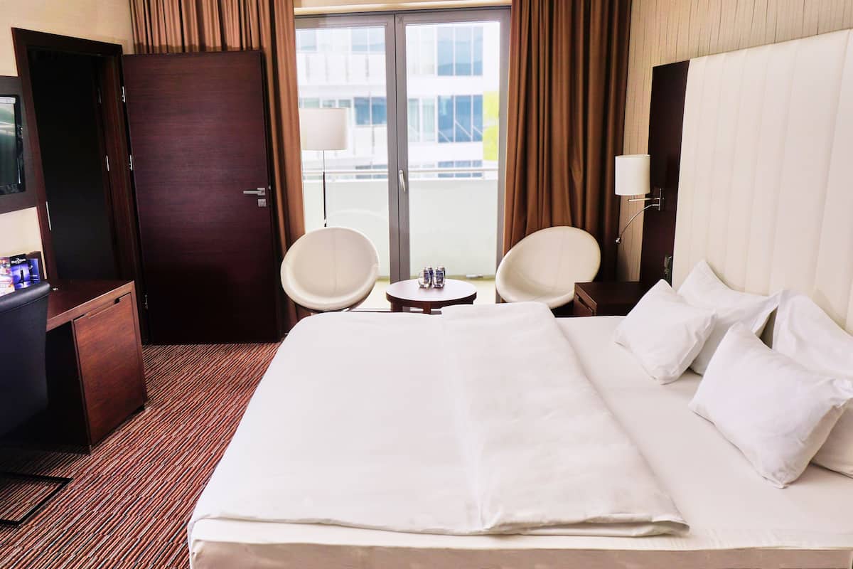Quarto de hotel com uma cama grande, duas cadeiras, uma mesinha, uma televisão e uma porta.