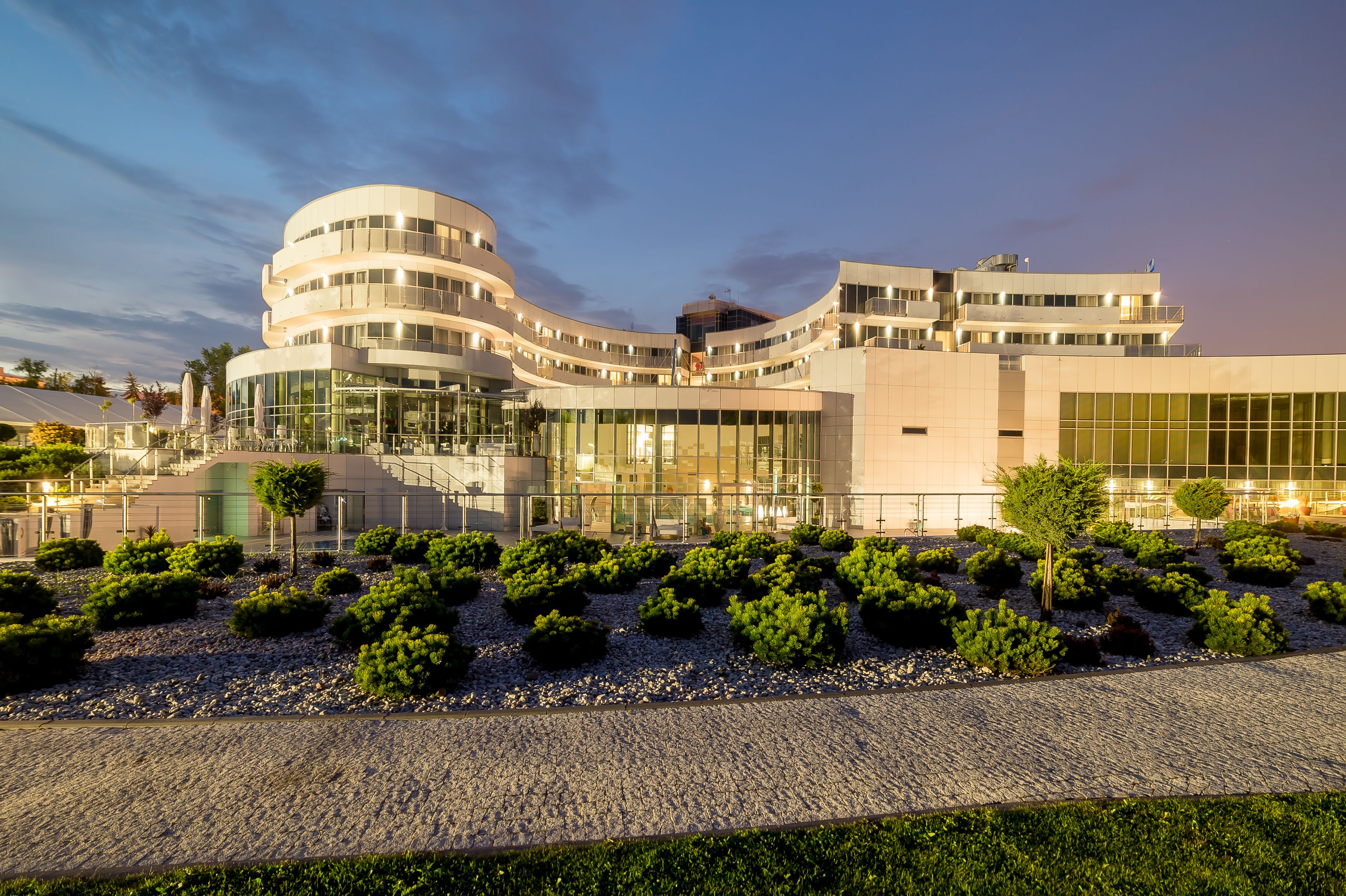 A modern building with a curved facade, multiple floors, and a landscaped garden with shrubs and trees.