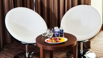 A hotel room with two white chairs, a small round table with a plate of fruit, a bottle of water, and a bottle of beer.