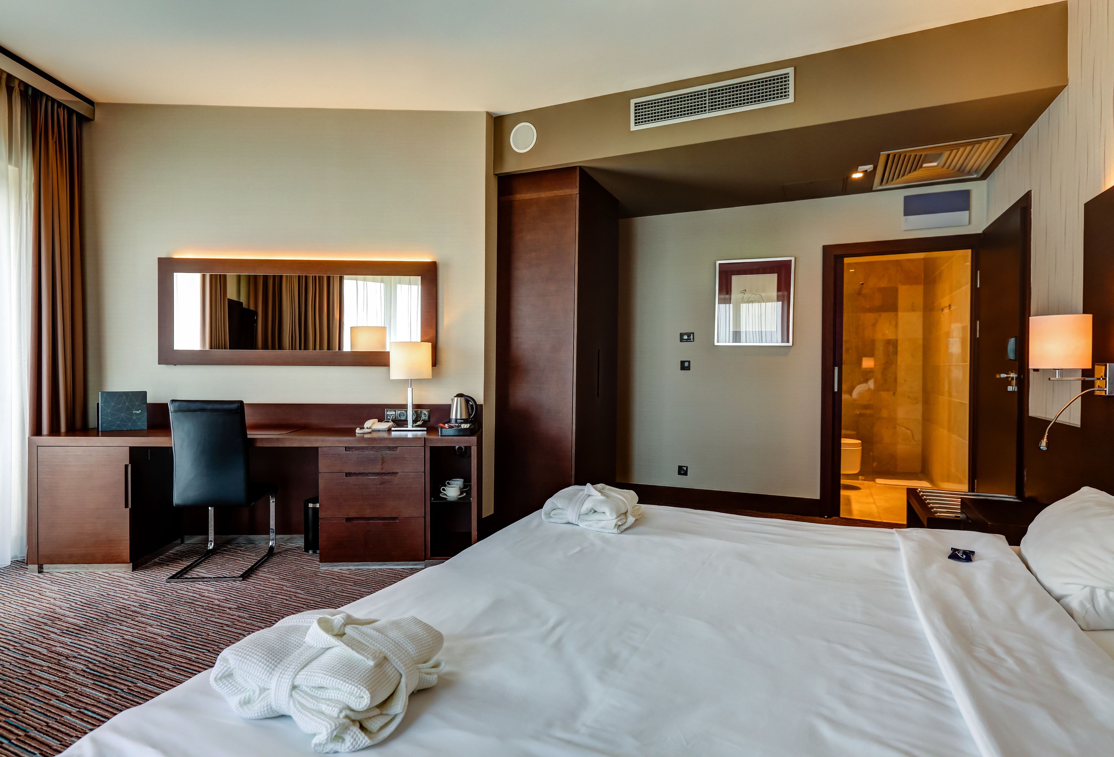 Hotelkamer met een groot bed, een bureau met stoel, een spiegel, een kledingkast en een zichtbare badkamer door een open deur.