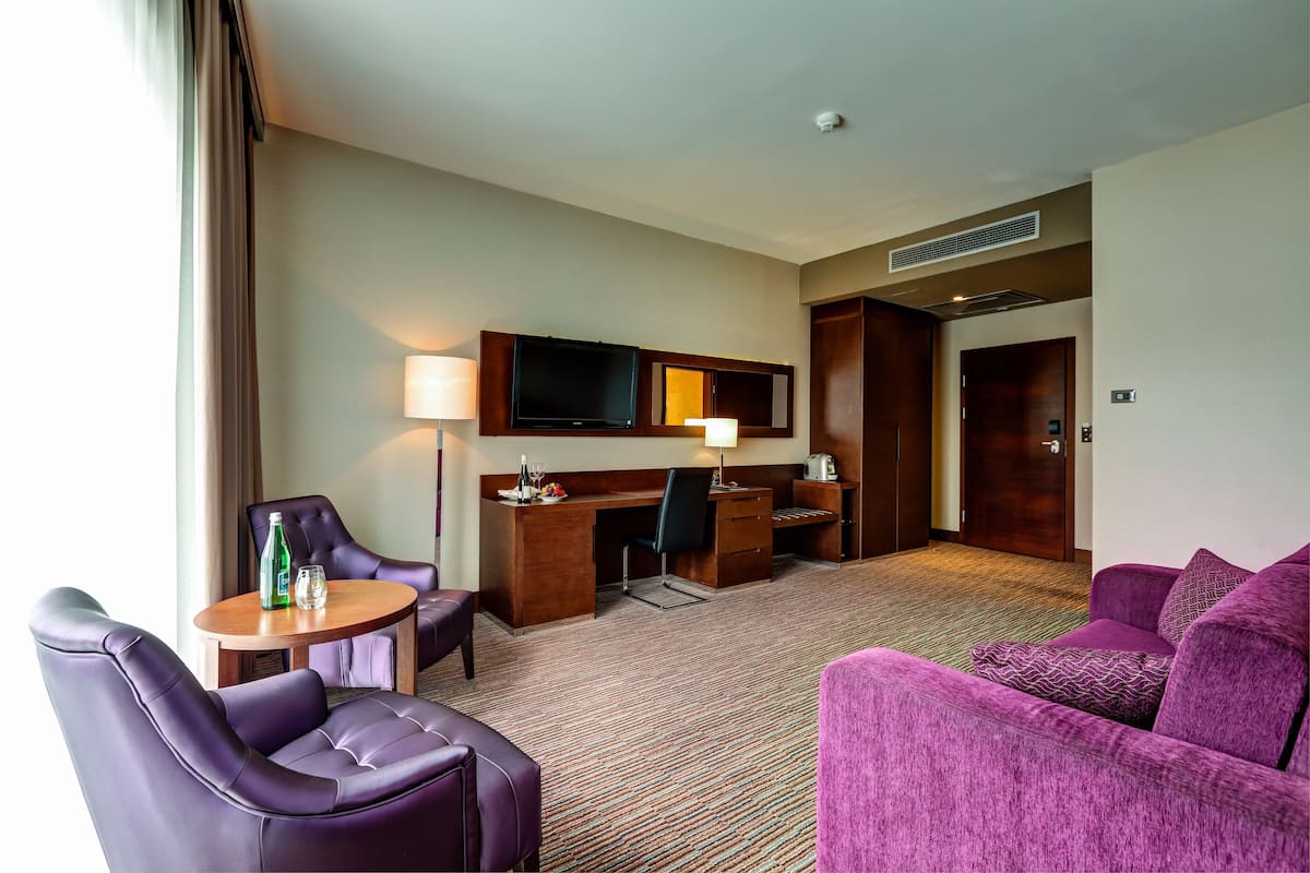 Quarto de hotel com sofá roxo, uma escrivaninha com cadeira, televisão e abajur.