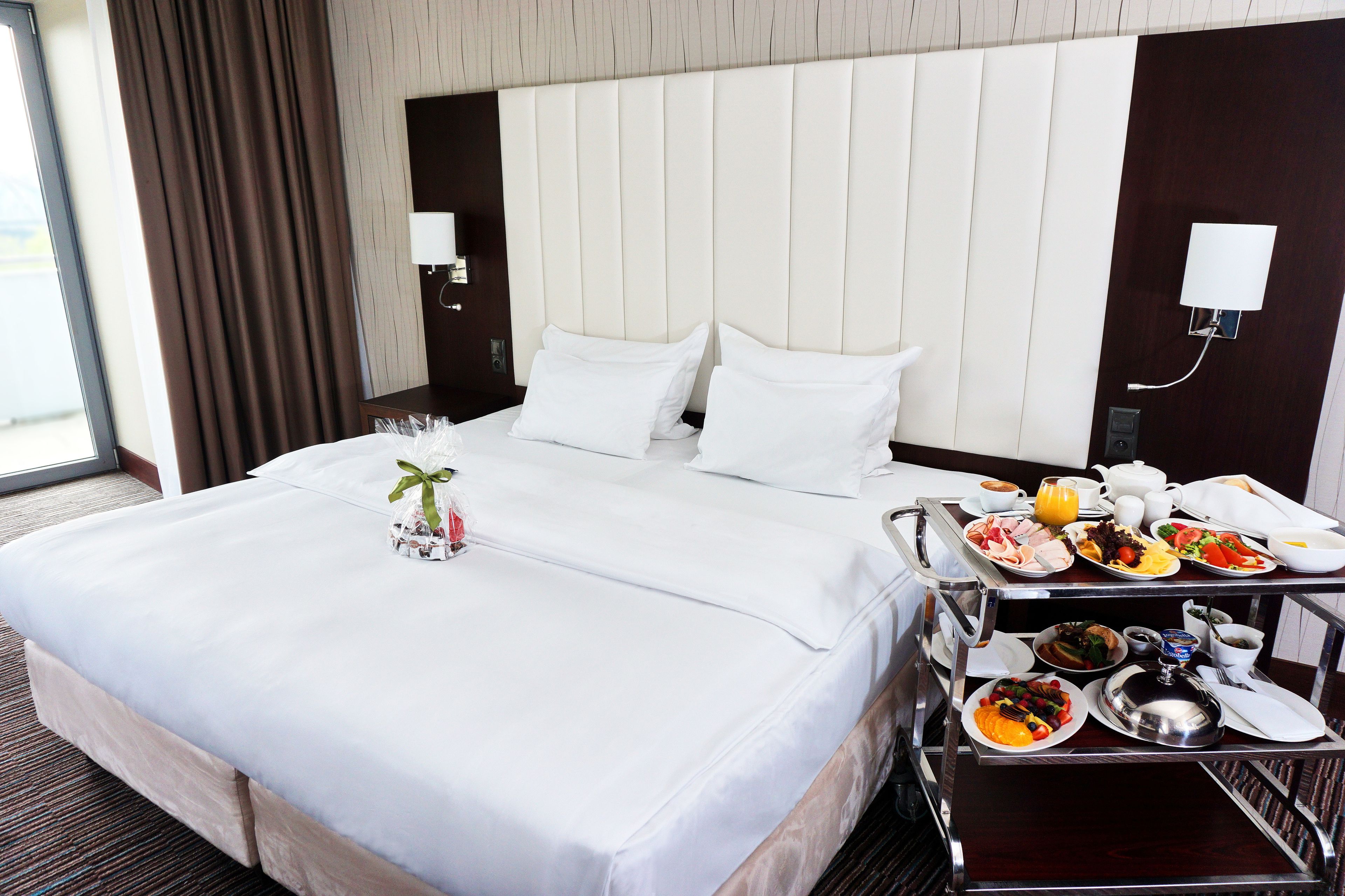 A hotel room with a large bed, a bedside table with a tray of breakfast items, and a nightstand with a lamp.
