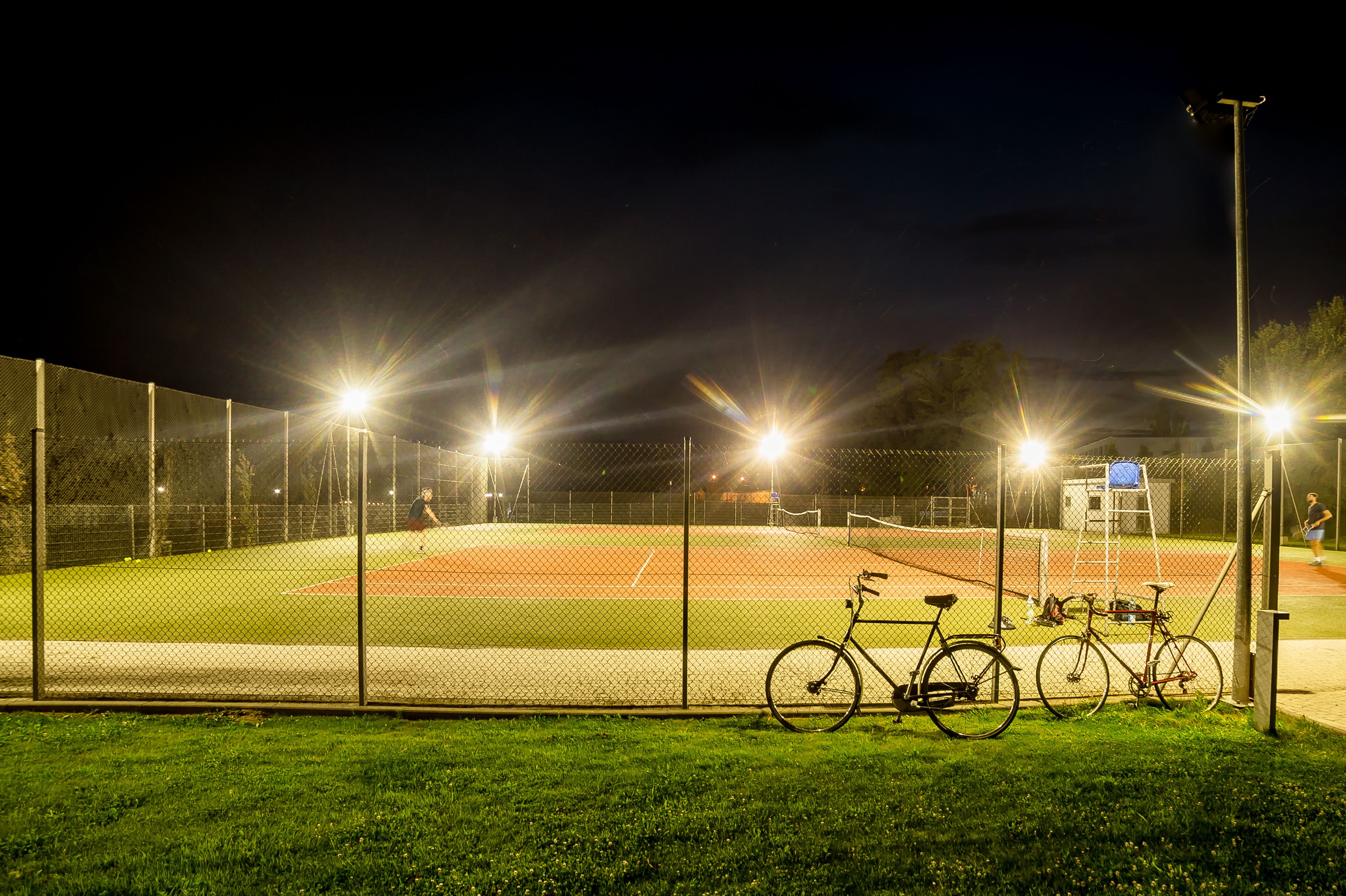 A baseball field at night with floodlights, a bicycle, and a fence.
