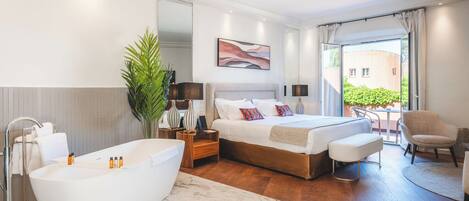 A modern bedroom with a large white bathtub, a double bed with a wooden headboard, a bedside table with lamps, and a view of the outside through a sliding glass door.