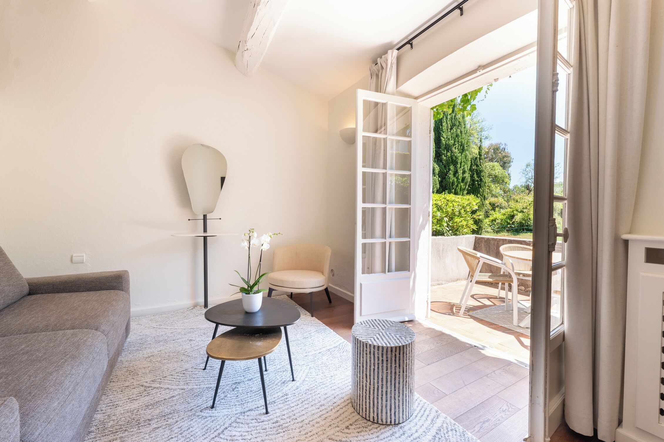 Un salon moderne avec un canapé, une petite table ronde agrémentée d’une plante, un lampadaire et une vue sur une terrasse extérieure avec une chaise et une table.