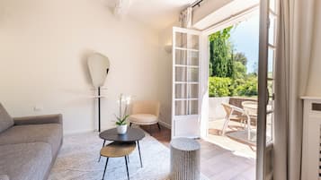 Un salon moderne avec un canapé, une petite table ronde agrémentée d’une plante, un lampadaire et une vue sur une terrasse extérieure avec une chaise et une table.
