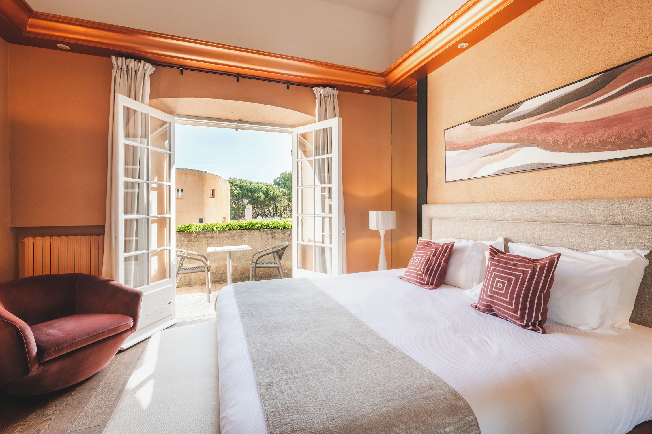 A hotel room with a large bed, a red armchair, a small table, and an open door leading to a balcony with a view of greenery.
