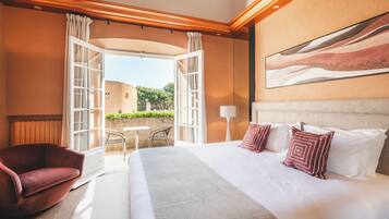 A hotel room with a large bed, a red armchair, a small table, and an open door leading to a balcony with a view of greenery.